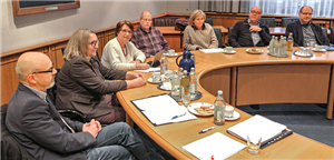 Vertreter/innen der Fraktion der Grünen im LWL und der Caritas in NRW sitzen an einem gebogenen Holztisch zusammen und reden miteinander