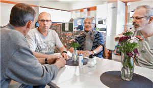 Drei Besucher und ein Sozialarbeiter sitzen im Café Emmaus in Rheydt an einem Tisch und reden miteinander. Im Hintergrund ist eine Küche mit zwei Frauen zu sehen.