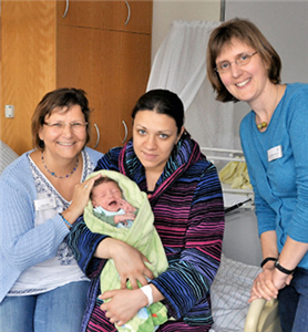 Zwei Frauen sitzen mit einem Baby auf einem Krankenbett. Eine weitere Frau steht daneben. Im Hintergrund sind ein Schrank und ein Kinderbett zu sehen.