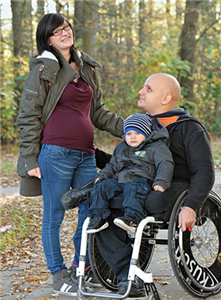 Ein junges Paar befindet sich mit ihrem Kind vor einem Wald. Die Schwangere steht neben ihrem, im Rollstuhl sitzenden, Mann, der ein kleines Kind auf dem Schoß sitzen hat.