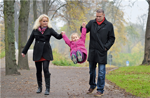 Ein junges Paar geht mit dem eigenen Kind durch einen Park spazieren. Das Mädchen hät sich an den Händen der Eltern fest und wird durch die Luft geschwungen.