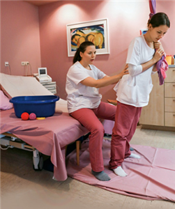 Zwei Hebammen befinden sich in einem Krankenzimmer und führen eine Übung durch. Dabei sitzt eine Hebamme auf einem Bett und lehnt die zweite sich an einem Seil festhaltende Hebamme nach vorne.