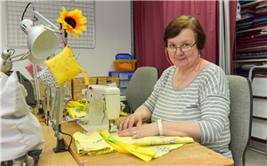 Eine ältere Frau sitzt an einem Arbeitsplatz vor einer Nähmaschine und stellt Produkte aus gelben, bedruckten Stoff her