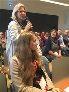 Eine Frau aus dem Publikum des Barcamps Soziale Arbeit am 25. und 26.10.2018 in Siegburg steht vor ihrem Stuhl und hält ein Mikrofon in der Hand.