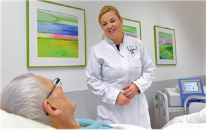 Eine Ärztin steht am Krankenbett eines Patienten und redet mit ihm. Im Hintergrund sind ein medizinisches Gerät und drei Bilder zu sehen.