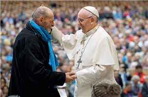 Ein Obdachloser steht mit Papst Franziskus beim Jubiläumsfest 'Fratello' in Rom vor Publikum auf einer Bühne. Der Papst hält die rechte Hand des Obdachlosen und streicht lächelnd über seinen Kopf.