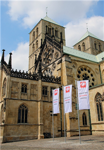 Der St.-Paulus-Dom in Münster aus der Seitenperspektiven. Neben dem Dom sind drei Fahnen-Masten mit Caritas-Flaggen in den Boden eingelassen