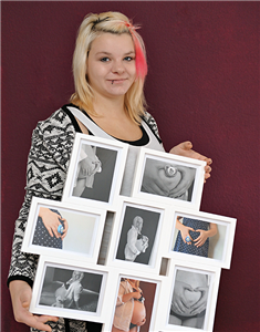 Eine junge Schwangere steht mit einem Galerie-Bilderrahmen vor einer dunkelroten Wand. In dem Rahmen sind Schwangerschaftsfotos von ihr zu sehen.