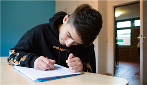 Ein Jugendlicher sitzt an einem Tisch und schreibt etwas auf ein Blatt Papier