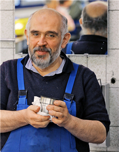 Ein Mann im Blaumann der ein Tuch und den Deckel einer Thermoskanne in den Händen hält. Im Hintergrund ist eine Wand mit grauen Fliesen, ein Spiegel und ein Seifenspender zu sehen.