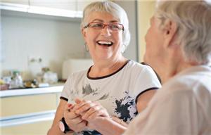 Eine Betreuerin sitzt lachend neben einer Seniorin und hält ihr die rechte Hand