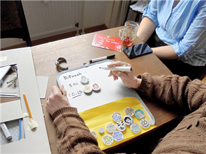 Eine Betreuerin und eine demente Seniorin sitzen an einem Holztisch. Vor der Seniorin liegt der Prototyp eine Tagesplaners, auf der sie ihre Planung mit einem Stift und Magneten vornimmt.