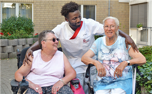 Zwei lächelnde Seniorinnen stehen, in ihrem Rollstuhl sitzend, in der Gartenanlage eines Altenzentrum. Ein dunkelhäutiger Pflege-Azubi steht hinter ihnen und legt seine Arme über ihre Schultern.