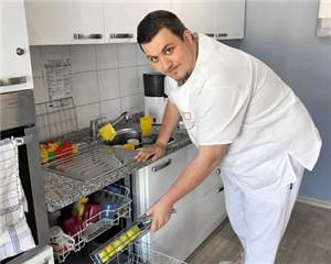 Ein FSJler räumt in einer kleinen Küche eine Spülmaschine aus und blickt dabei in die Kamera
