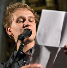 Ein junger Mann steht vor einem Stehmikrofon mit einem Zettel in der Hand und trägt seinen Beitrag zum Poetry-Slam auf der 'Pflege-Gala' am 12. Mai 2019 in Paderborn vor