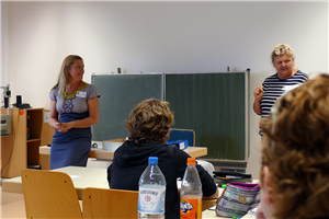 Menka Berres-Förster steht an einem Pult in einem Klassenzimmer und beobachtet eine Lehrerin und einige Schüler beim Unterricht