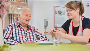 Eine Pflegerin sitzt mit einem Senior an einem Tisch und schält einen Pfirsich. Der Senior schaut ihr dabei zu.
