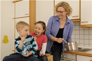 Eine Frau steht mit einem Kochtopf in einer Küche. Daneben sitzen ein Junge und ein Mädchen, die beide jeweils einen Joghurt essen.