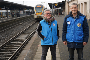 Eine Mitarbeiterin und ein Mitarbeiter der Bahnhofsmission stehen auf einem Gleis des Paderborner Hauptbahnhofs. Im Hintergrund ist ein stehender Zug zu sehen.