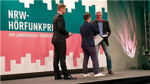 Christian Heine-Göttelmann steht mit dem Preisträger des Sozialpreises Florian Rübener auf der Bühne des LfM-Hörfunkpreises 2019 und beglückwünscht ihn. Der Moderator der Veranstaltung steht daneben.