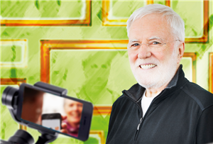Porträt: Gerd Fischer, einem hell-grünen Hintergrund mit goldenen Bilderrahmen. Im Vordergrund ist ein Smartphone auf einem Selfie-Stick zu sehen.