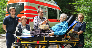 Ein Malteser-Herzenswunsch-Krankenwagen, der vor einem Wald steht. Neben dem Wagen steht eine Krankenbahre mit einem Patient und vier Helfer/innen.