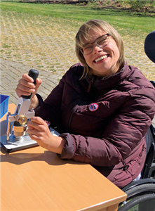 Eine Beschäftigte der CBW in Würselen, die in ihrem Rollstuhl im Außenbereich an einem Tisch sitzt und an einer Buttonmaschinen kleine Button zur Distanzregelung herstellt