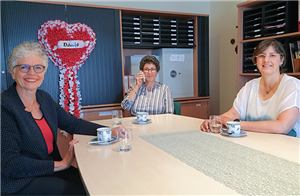 Marion Peters, Evelyn von Heel und Kerstin Mengeler sitzen zusammen an einem Konferenztisch. Im Hintergrund sind Schränke mit einem Tresor, vielen Ablagekörbchen und ein dekoratives Herz zu sehen.