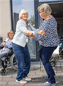 Zwei Frauen, die sich auf der Terasse eines Altenheims befinden, tanzen miteinander. Im Hintergrund sitzt eine weitere Frau in einem Rollstuhl.