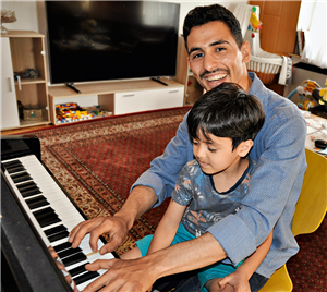 Aeham Ahmad sitzt in seiner Wohnung an einem Klavier, spielt und blickt dabei lächelnd in die Kamera. Sein Sohn sitzt dabei auf seinem Schoß.