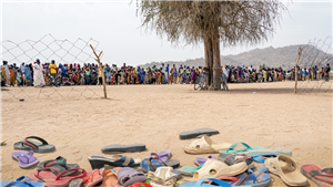Ein Flüchtlingslager in Igawa im Norden Kamerums: Im Hintergrund ist eine große Menschenmenge zu sehen, im Vordergrund sind diverse Schuhe aufgehäuft. Dazwischen sind Drahtzäune und eine Baum zu sehen