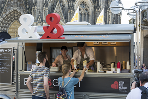 Der Imbisswagen der Aktion 'Eat & Greet', der auf der Kölner Domplatte steht. In dem Wagen arbeiten zwei Mitarbeitende, die eine Frau und einen Mann bedienen.