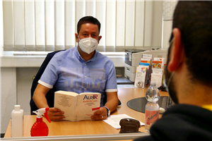 Samer Qarqash sitzt mit Schutzmaske und einem aufgeschlagenen Buch zum Ausländerecht in einem Büro an einem Schreibtisch und guckt einen Klienten mit Schutzmaske an, der ihm gegenüber sitzt