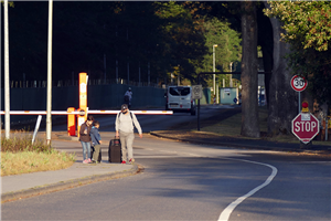 Zufahrtsstraße zur EAE Mönchengladbach mit einigen Verkehrsschildern und einer Schranke. Auf der Straße fährt ein weißer Lieferwagen, auf dem Bürgersteig geht ein Geflüchteter mit zwei Kindern.