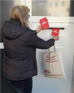 Eine Ehrenamtliche steht vor einem Wohnhaus und wird Postkarten in einen Briefkasten. Sie trägt dabei einen Beutel über dem Arm.