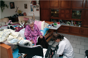 Eine Seniorin sitzt auf einem Pflegebett, das in einem Wohnzimmer steht, und wird von einer Pflegerin, die vor ihr kniet, betreut