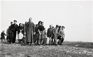 Eine schwarz-weiß Aufnahme mit einer Gruppe aus Kriegsflüchtlingen, die mit ihren Habseligkeiten über ein Feld läuft. Die Gruppe besteht zu großen Teil aus Kindern und Seniorinnen.