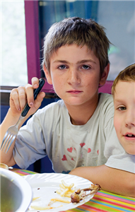 Ein Junge sitzt vor einem leeren Teller und hält eine Gabel in der Hand. Neben ihm sitzt ein weiterer Junge.