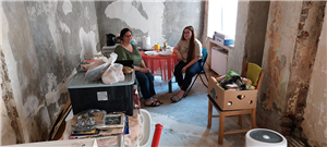 Zwei Frauen sitzen in einem Raum, der aufgrund von Hochwasserschäden entkernt wurde, zwischen einigen Möbeln an einem Tisch und blicken in die Kamera