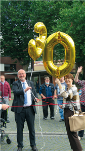 Der Paderborner Diözesan-Caritasdirektor Josef Lüttig hält zusammen mit einer Frau zwei goldenen Luftballon, die die Zahl 30 darstellen, in der Hand. Zwischen ihnen wurde eine rote Schnur gespannt.