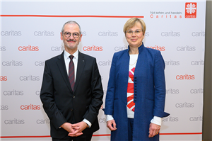 Peter Neher und Eva M. Welskop-Deffaa stehen zusammen vor einer Stellwand der Caritas und lächeln in die Kamera