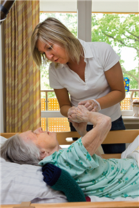 Eine Pflegerin steht an einem Krankenbett, in dem eine Seniorin liegt. Beide halten sich an den Händen und blicken sich in die Augen.