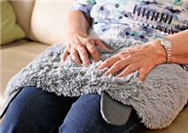 Eine Seniorin, die auf einem hellen Sofa sitzt. Auf ihrem Schoß liegt ein hellgraues Fell, auf das sie ihre Hände ablegt.