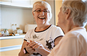 Eine weibliche Betreuungskraft sitzt mit einer Seniorin in einer Küche, hält ihre Hand und lächelt
