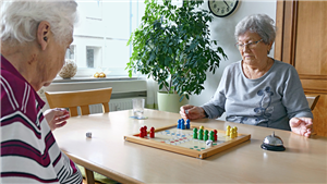 Zwei Seniorinnen sitzen bei einer Tagespflege in einem Gemeinschaftsraum an einem Tisch und spielen "Mensch ärgere dich nicht" 