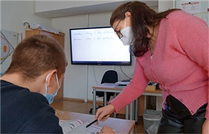 Eine Mitarbeiterin der Einrichtung "lern.punkt" steht neben einem Schüler in einem Klassenraum und deutet mit einem Stift auf eine Stelle in einem Lehrbuch