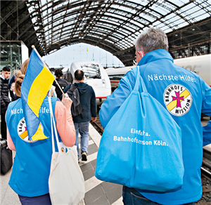 Zwei Mitarbeitende der Bahnhofsmission Köln, die mit Hilfsgütern über einen vollen Bahnsteig des Kölner Hauptbahnhofs laufen