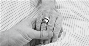 Die Hand einer Seniorin mit einigen Ringen am Ringfinger, die auf einer Bettdecke liegt und von einer anderen Hand gehalten wird