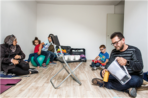 Eine Familie mit drei Kindern sitzt in einem nicht möblierten Raum mit ein wenig Gepäck auf dem Boden. In der Mitte steht ein Klappstuhl, auf dem ein Tablet mit Bechern steht.
