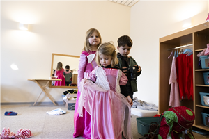 Zwei Mädchen und ein Junge stehen zusammen in einem Ankleidezimmer einer Kita. Die Mädchen tragen ein pinkes Kleid, der Junge ist als Fotograf verkleidet.